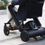 Close-up of the lower half of a person seated in a black Whill Model F power wheelchair, focusing on the chair's base and wheels.