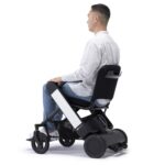 A person in a white shirt and jeans seated in a white armrests Whill Model F power wheelchair, viewed from the side.