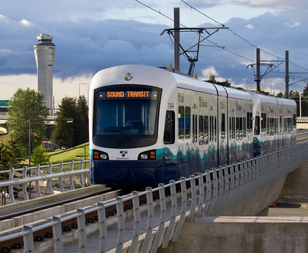 A Sound Transit light rail train on an elevated track near Seattle's airport, with the control tower in the background.