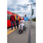 A senior woman riding the eFOLDi Lite mobility scooter on a train platform, showcasing the scooter's utility in public transportation settings, with a family walking alongside.