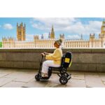 An elderly woman enjoys a sunny day, riding on the eFOLDi Lite mobility scooter with the iconic Houses of Parliament in the background, symbolizing freedom and mobility.