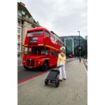 A stylish elderly woman stands on a city sidewalk with the eFOLDi Lite mobility scooter in its compact, folded form next to her, as a classic red double-decker bus passes by in the background.