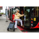 A woman stepping onto a red city bus while pulling the black eFOLDi Lite mobility scooter behind her, which is folded into a compact size resembling carry-on luggage.