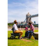 An individual seated on the eFOLDi Explorer mobility scooter, conversing with a person on a bench in a park with a famous bridge in the background, illustrating the scooter's use in recreational areas.