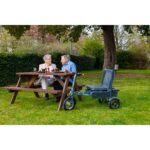 An elderly couple sit at a garden table, exuding a sense of calm and companionship. Beside the man stands the eFOLDi Explorer Foldable Ultra Lightweight Mobility Scooter, blending seamlessly into the tranquil outdoor setting, symbolizing ease of mobility and quality of life.