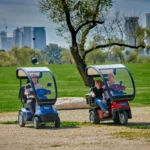 Live Style Image Side View of Blue Afiscooter S4 with Canopy Mobility Scooter, Red Afiscooter S3 Dual Seat Mobility Scooter with Canopy Golf Tires