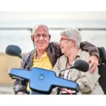 An elderly couple shares a joyful moment on a blue Afiscooter S Dual Seat mobility scooter, smiling and laughing together, with the man affectionately holding the woman close.