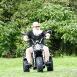 Man riding black Daymak Boomerbeast 2D on grass with green trees.