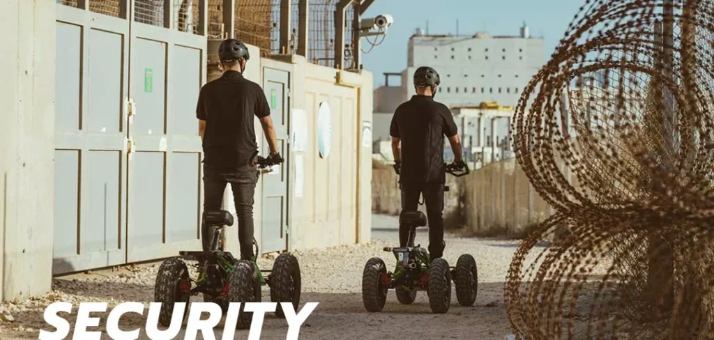 Two security officers wearing helmets patrolling a fenced area on EZRaider HD4 4x4 off-road electric scooters near barbed wire and industrial buildings