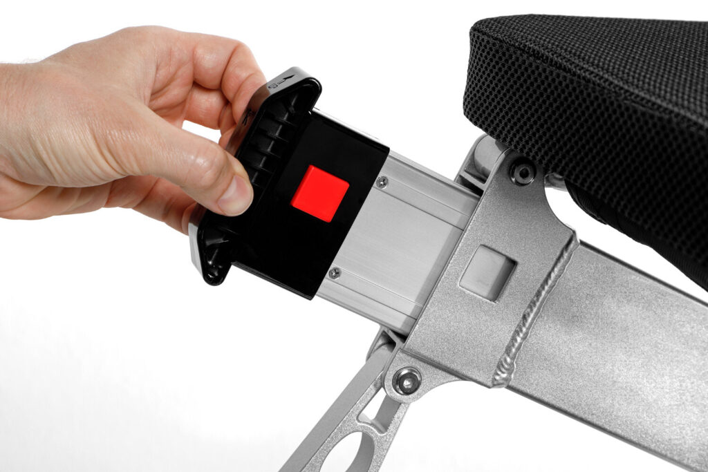 Porto Mobility Ranger Discovery XL folding power wheelchair, hand holding a pack with red button at the end of the frame tube below the seat